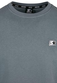 Grauer Baumwoll-Sweatshirt mit Rundhalsausschnitt, das auf der linken Brustseite ein kleines, gesticktes Logo in Weiß und Schwarz zeigt. Glatte Textur.