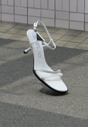 White strappy high-heeled sandal with a thin heel, black sole, and brand name "Replay" on the insole, photographed on asphalt near tiled wall.
