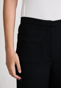 Black textured trousers with a wide waistband and a side pocket, paired with a plain white top. Smooth fabric finish.
