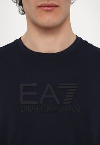 Tamnomodra bombažna majica s kratkimi rokavi, ki na prsih vsebuje dvignjen črn logotip EA7 Emporio Armani. Ima okrogel izrez.