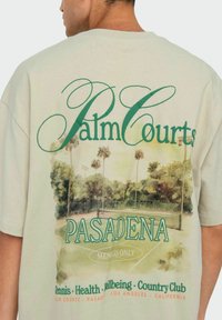 Hellbeige übergroßes T-Shirt mit einem grünen und orangefarbenen Grafikdesign, das einen Tennisplatz, Palmen und den Text "Palm Courts Pasadena" zeigt.