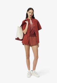 Camisa roja de botones, pantalones cortos a juego y un bolso crema con acento marón. Zapatillas blancas completan el atuendo. Un look casual y coordinado.