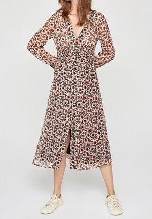 Femme portant une robe midi à manches longues fleurie avec une fente à l’avant et des baskets blanches décontractées sur un fond uni.