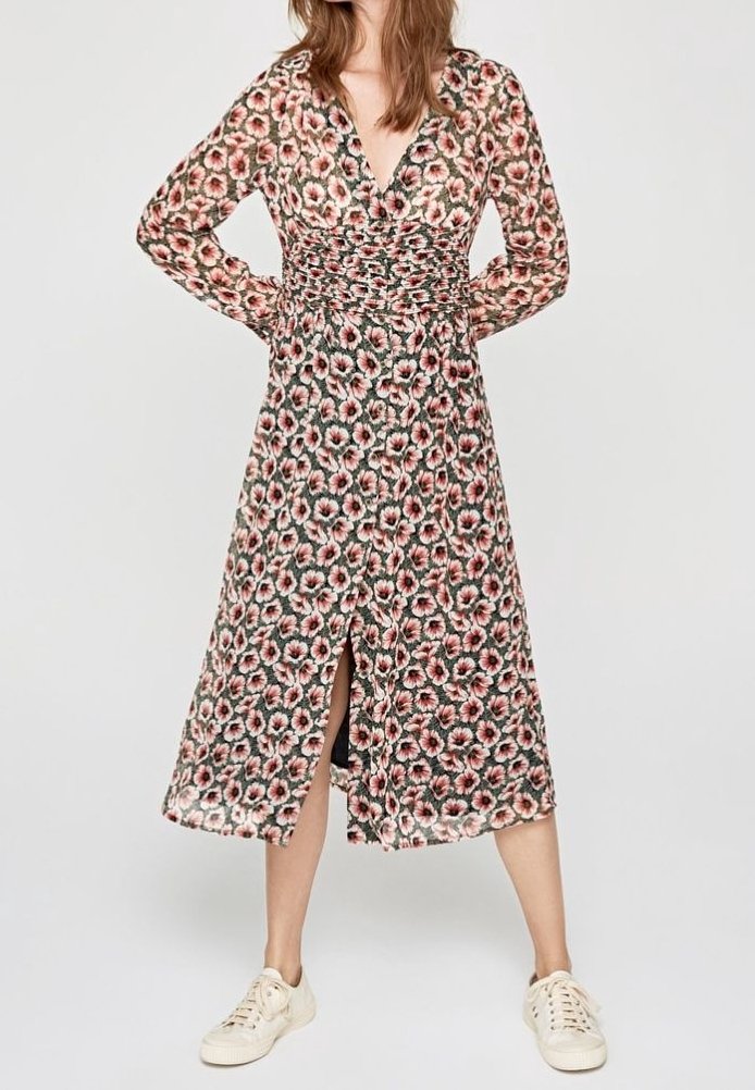Femme portant une robe midi à manches longues fleurie avec une fente à l’avant et des baskets blanches décontractées sur un fond uni.