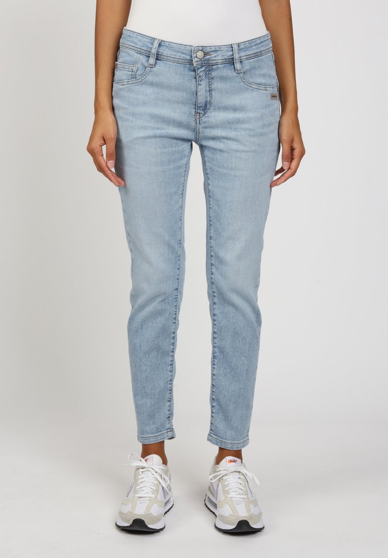 Hellblaue Jeans aus Denim mit schmaler Passform, ausgestattet mit Taschen, einem Knopfverschluss und dezentem Verblassen; kombiniert mit weißen und grauen Turnschuhen.