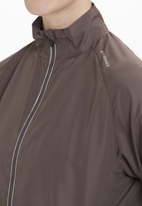 Brun lättviktsjacka med hög krage, frontdragkedja och reflexdetaljer på bröstet. Materialet verkar slätt och vattentåligt.