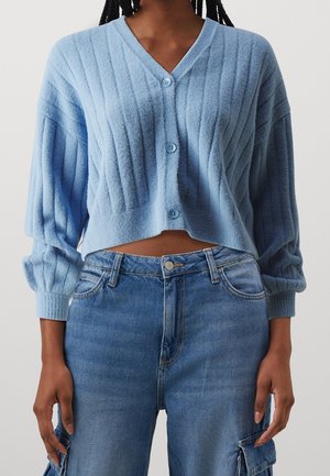 Femme portant un cardigan court côtelé bleu clair avec des boutons et un pantalon cargo en denim taille haute sur un fond uni.