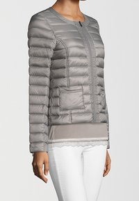 Femme portant une veste matelassée gris ajustée avec une fermeture éclair à l'avant, des poches plaquées et un pantalon blanc sur un fond uni.