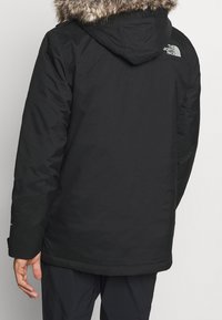 Veste isolante noire avec une capuche en fourrure douce, dotée d'un extérieur imperméable et d'un logo dans le dos, conçue pour les temps froids.