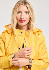Veste imperméable jaune avec capuche, dotée d'une fermeture éclair frontale, de boutons-pression et de cordons de tirage ajustables. Poignet à motifs visible.