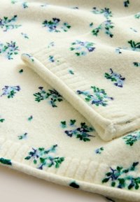 Kreemikas lillemustriga fliisitekik, millel on sinised ja rohelised lilled, pehme tekstuur, volditud nurk, mis paljastab ribakujulise aktsendi ääres.