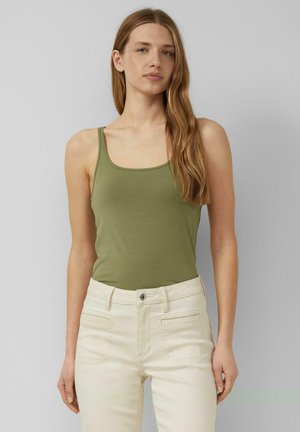 Kobieta z długimi jasnobrązowymi włosami, ubrana w oliwkowo-zieloną koszulkę na ramiączkach i białe spodnie z wysokim stanem na szarym tle.