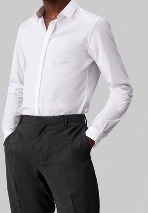 Homme portant une chemise blanche cintrée rentrée dans un pantalon gris foncé taille haute, ajusté, avec les mains dans les poches.