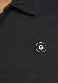 Svart polo-shirt med en texturerad tyg. Har en cirkulär broderad logotyp i svart och vitt på vänster bröst.