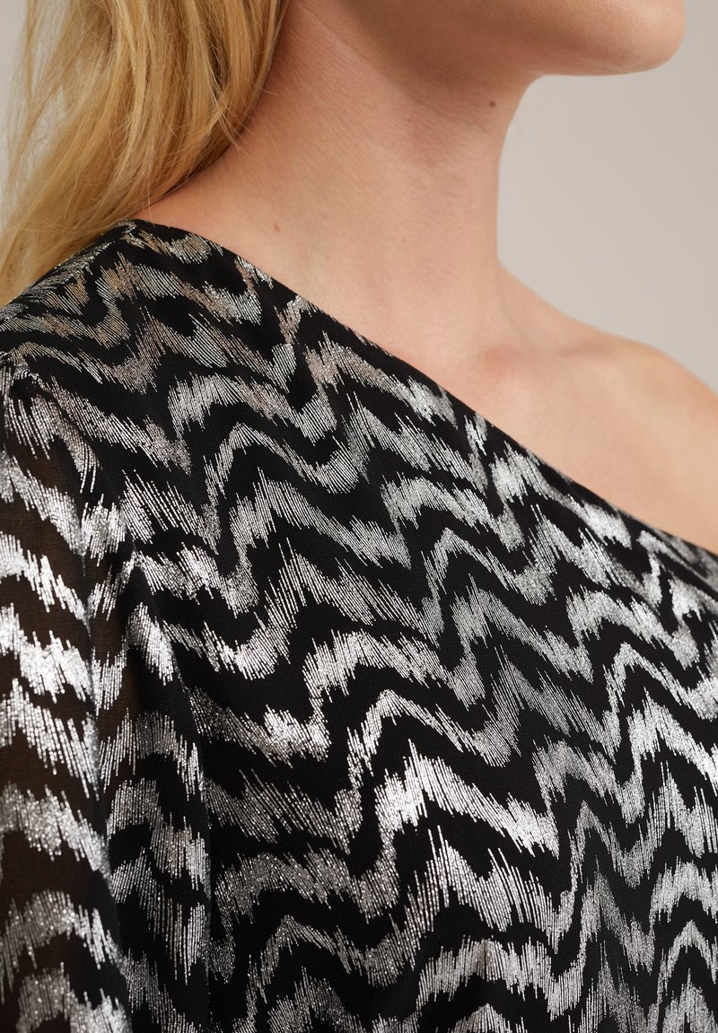 Schwarzer Stoff mit einem Zickzackmuster aus silbernem Faden, der über eine Schulter fällt, mit einer weichen, strukturierten Optik.