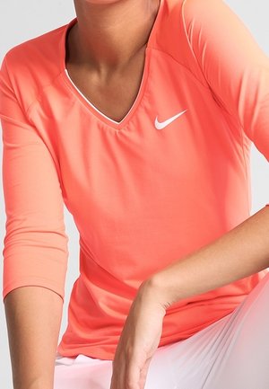 Personne portant un haut à manches longues corail de Nike avec un col en V et un pantalon blanc, assise avec un bras reposant sur son genou.