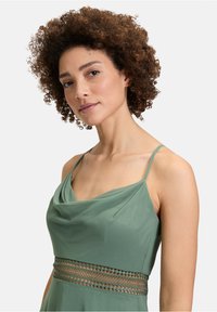 Sukienka z dekoltem typu "cowl" w miękkim, stłumionym zielonym materiale, z cienkimi ramiączkami i ozdobnym akcentem koronkowym w talii. Gładka tekstura i luźny kroj.