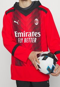 Rotes Fußballtrikot mit Zickzack-Muster, schwarzen Akzenten und Kapuze. Enthält ein Logo, Markennamen und wird mit einem schwarz-weißen Fußball kombiniert.