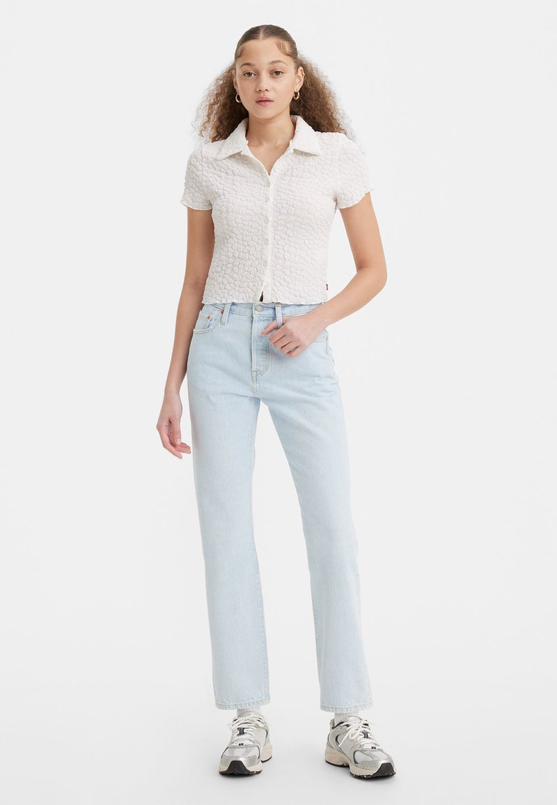Chemise blanche à texture et à manches courtes avec un design à boutons, associée à un jean droit bleu clair et des baskets grises.