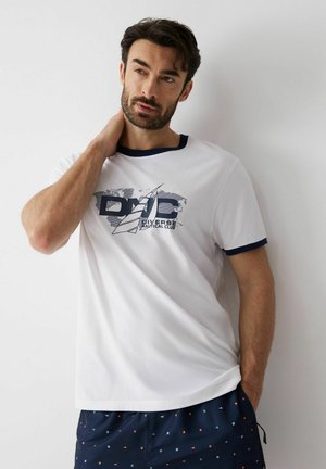 Mężczyzna w białej koszulce z logo klubu żeglarskiego i granatowych szortach z małymi kolorowymi wzorami, stojący przy białej ścianie.