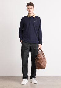 Polo a maniche lunghe blu navy con colletto beige, abbinato a pantaloni in denim scuri e sneakers bianche. Borsa in pelle marrone tenuta nella mano destra.