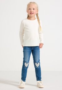 Malá dievčina s blond vrkočmi, oblečená v bielej košeli s dlhým rukávom, modrých džínsoch s nášivkami srdca na kolenách a bielych teniskách, stojí na bielom pozadí.