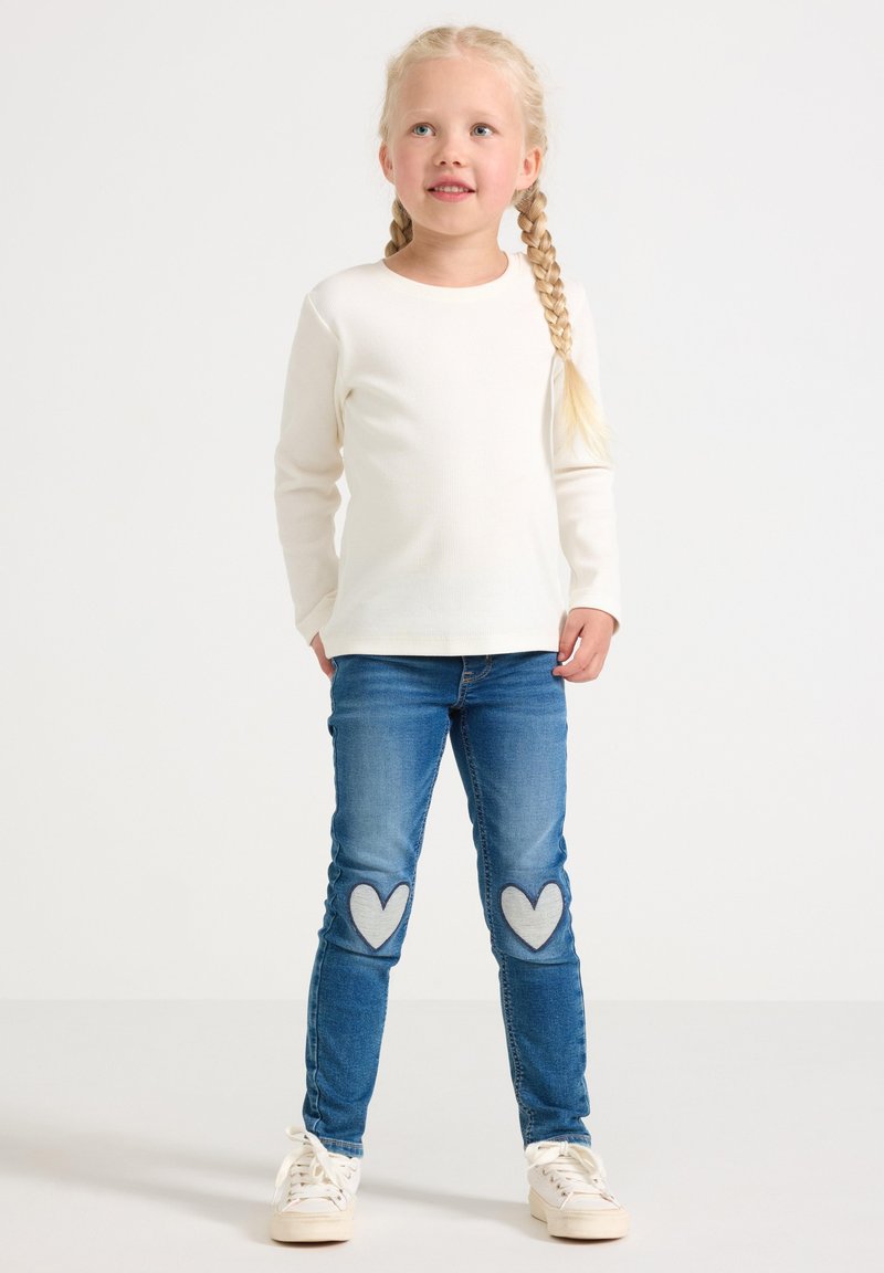 Malá dievčina s blond vrkočmi, oblečená v bielej košeli s dlhým rukávom, modrých džínsoch s nášivkami srdca na kolenách a bielych teniskách, stojí na bielom pozadí.