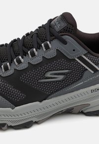 Szare buty sportowe z siateczkową cholewką, gumową podeszwą i wzmocnionymi akcentami. Posiadają logo, teksturowane wzory oraz kontrastowe sznurowadła i nałożone elementy.