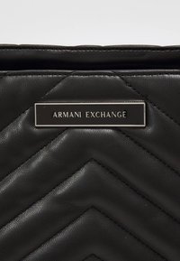 Bolso negro de piel sintética acolchada con una placa prominente con el logo plateado de "Armani Exchange". Patrón en espiga y textura lisa.