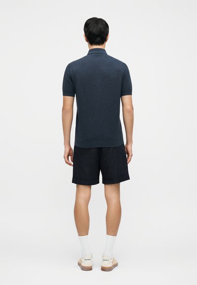 Σκούρο μπλε πουκάμισο πόλο με κοντά μανίκια και γιακά· συνοδεύεται από σκούρα σορτς· το μοντέλο στέκεται με την πλάτη στην κάμερα, με άσπρες κάλτσες και ελαφριά παπούτσια.
