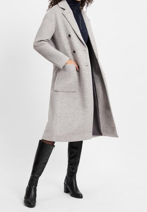 Vrouw die een lichtgrijze, knielange jas met grote zakken draagt, zwarte hoge laarzen met hakken en een donker binnenoutfit.