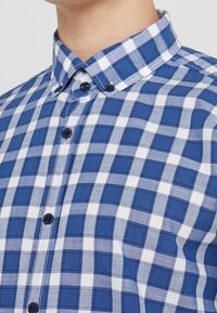 Camicia a quadri blu e bianca con colletto, caratterizzata da bottoni scuri e un tessuto tessurizzato. Il motivo è composto da ampi quadrati a scacchi.