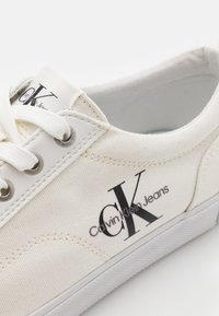 Vit canvas sneakers med snörning, rund tå och en stor svart logotyp med texten "Calvin Klein Jeans" på sidan.