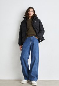 Sort polstret jakke med hætte, grøn sweater under, løse blå denimjeans og hvide sneakers. Bløde og kontrasterende teksturer.