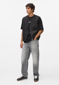 Zwarte gewatteerde bodywarmer met ritsluiting over een zwart T-shirt, gecombineerd met lichtgrijze jeans en zwarte sneakers met witte accenten.