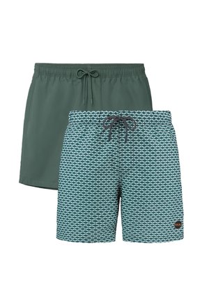 Jonge man met krullend haar, blootsvoets staand, met turquoise zwemshorts, armen gekruist, tegen een effen witte achtergrond.
