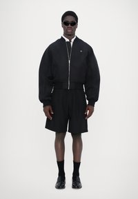 Veste bomber noire courte avec fermeture éclair, associée à un short noir. Le modèle porte des lunettes de soleil noires et des mocassins noirs avec des chaussettes de longueur mi-mollet.