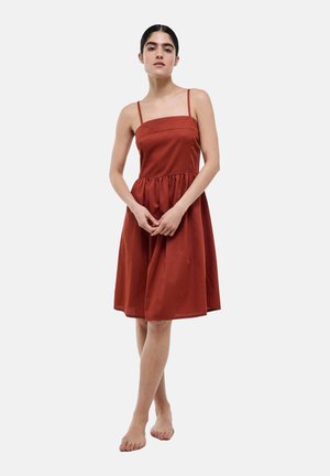 Femme debout pieds nus portant une robe sans manches couleur rouille, longueur genoux, avec fines bretelles sur un fond blanc uni.