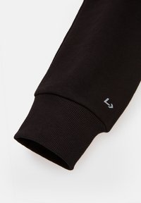 Manche de sweat-shirt noire avec poignet côtelé et petit logo réfléchissant près du poignet sur fond blanc uni.