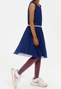 Abito senza maniche blu navy con gonna arricciata e cintura con accento dorato, abbinato a leggings lucidi viola e sneaker bianche con dettagli pastello.