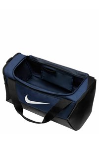 Sac de sport Nike en bleu foncé avec un logo blanc, comprenant un compartiment principal zippé, un fond noir et des poignées doubles pour un transport facile.