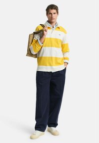 Langdurige poloshirt met gele en witte strepen, gecombineerd met marineblauwe broek. Accessoires omvatten een tan totebag en lichte sneakers.