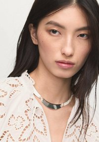 Sølv choker halskæde med en buet metalplade, der har hvide led. Modellen bærer en hvid blondebluse med indviklede mønstre.