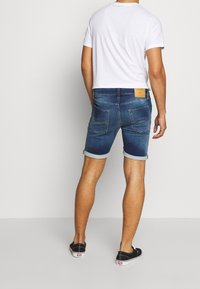 Bermudas de mezclilla azul para hombre con dobladillos enrollados, fabricadas en algodón, con dos bolsillos traseros y una etiqueta de cuero marrón en la cintura. Llevadas con una camiseta blanca.