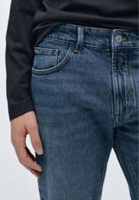 Modré džíny z denimu s rovným střihem, pěti kapsami, jemným vyblednutím a kontrastním prošitím. Ruka spočívající na bok.