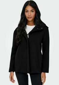 Veste noire zippée avec capuche, fabriquée en tissu doux. Présente un design structuré, des manches longues et des détails en métal à pression sur le col.