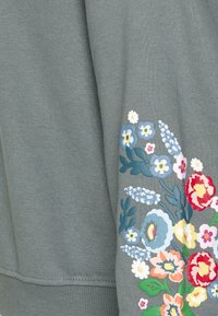 Sweatshirt vert ornée de broderies florales vibrantes en bleu, rose, rouge et jaune sur la manche droite. Tissu doux et texturé.