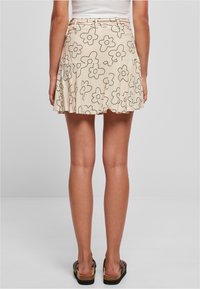 Jupe courte beige avec un motif de fleurs noires, design plissé et ceinture. Présentée de dos, portée avec des sandales plates.