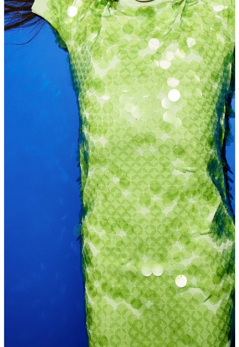 Gros plan d'une personne portant une robe verte ornée de grands sequins circulaires contre un fond bleu uni.