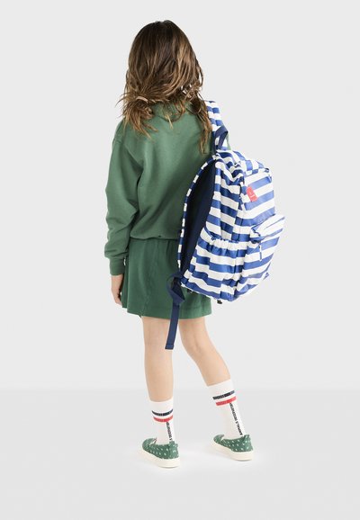 Zaino a righe blu e bianche con dettagli arricciati; felpa verde e gonna; calzini bianchi con righe rosse e nere; scarpe verdi a pois.